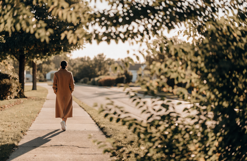A woman in a camel coat walking a tree-lined sidewalk at golden hour